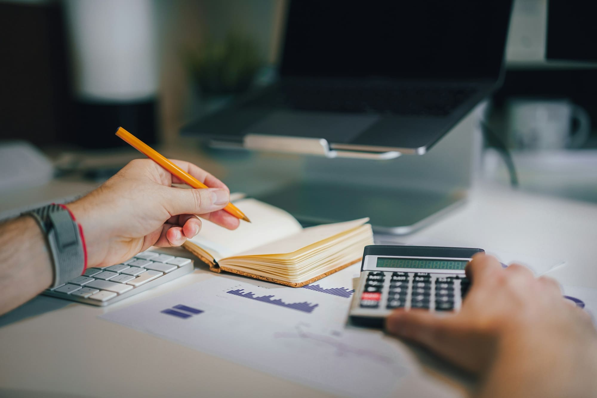 Hands work with a calculator, pencil and notebook over printed charts.