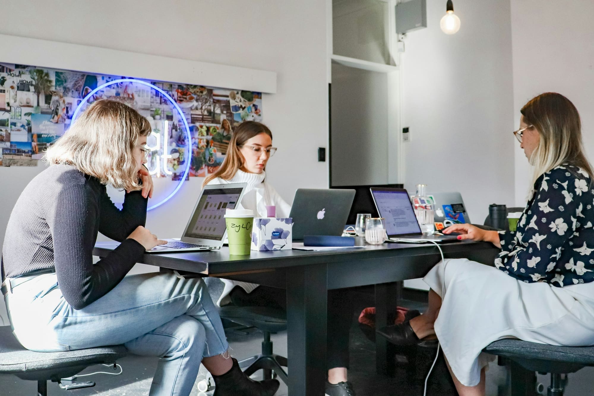 Three women work on laptops at a table in a bright studio with a photo wall.