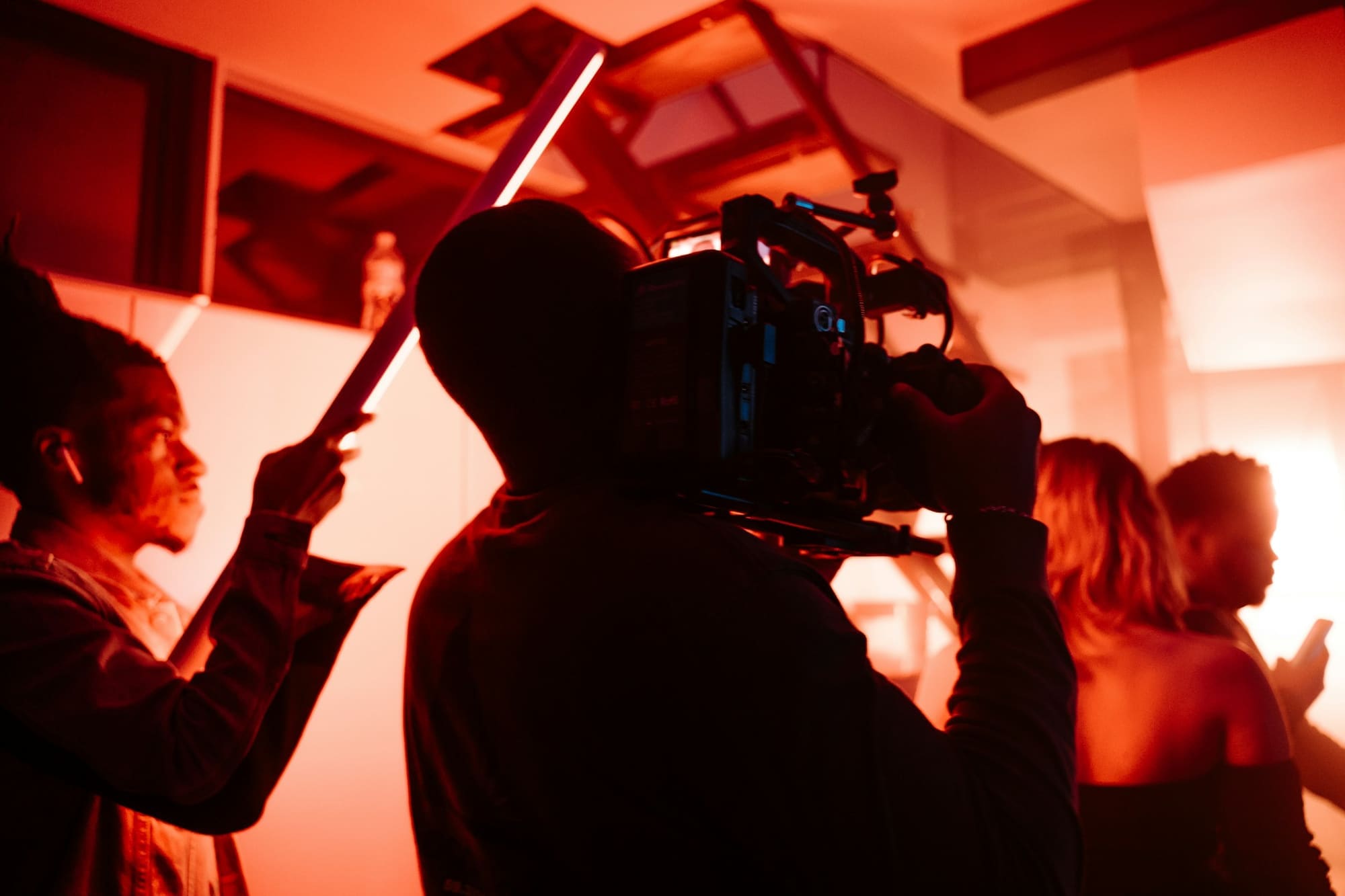A film crew shoots in red light: a camera operator shoulders a cinema camera while a crew member holds a light tube.