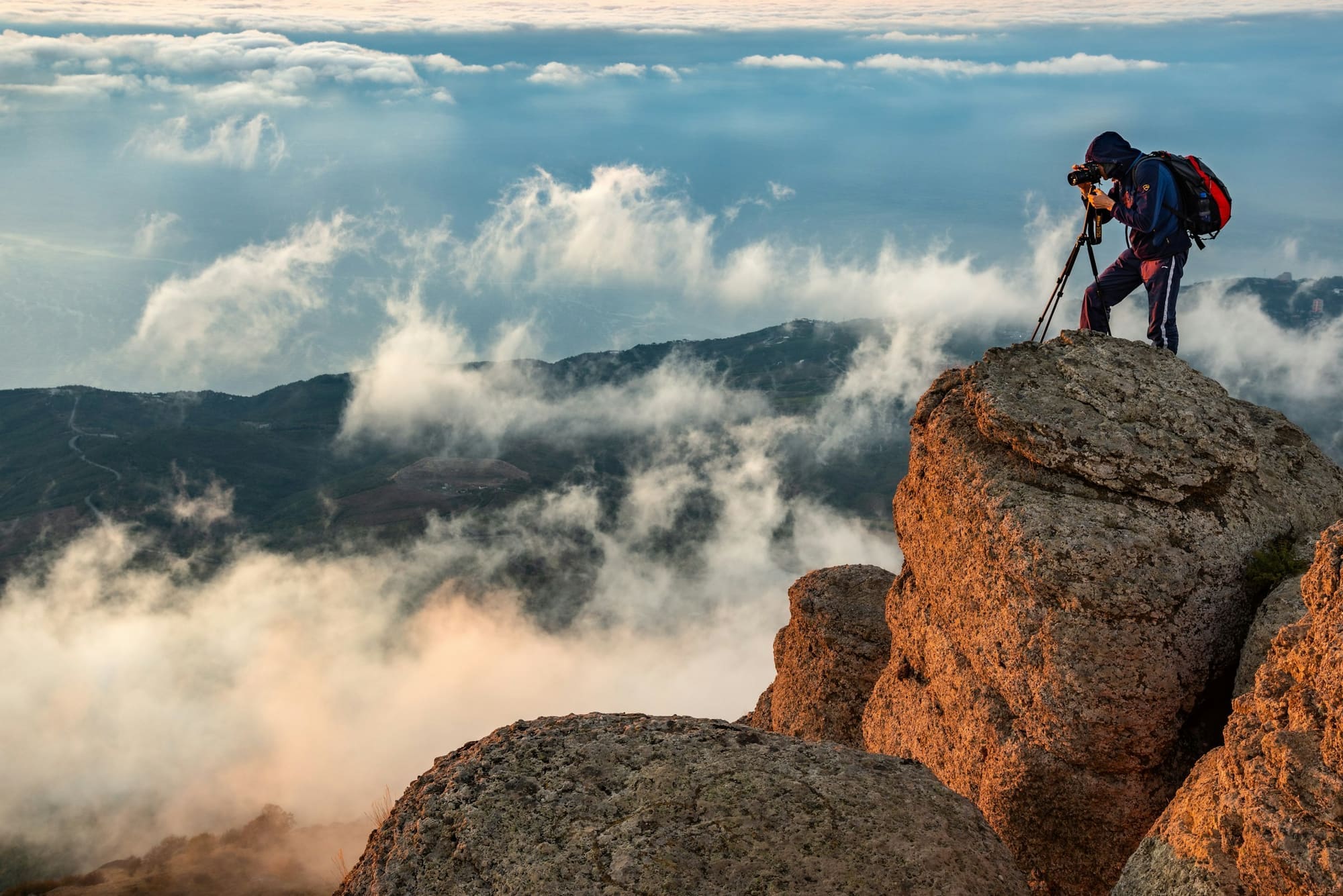 A photographer with a tripod stands on a rocky summit above a sea of clouds.