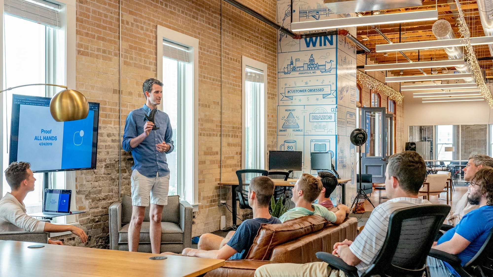 A presenter speaks to a seated team in a brick-walled loft office, with a slide on a screen behind him.