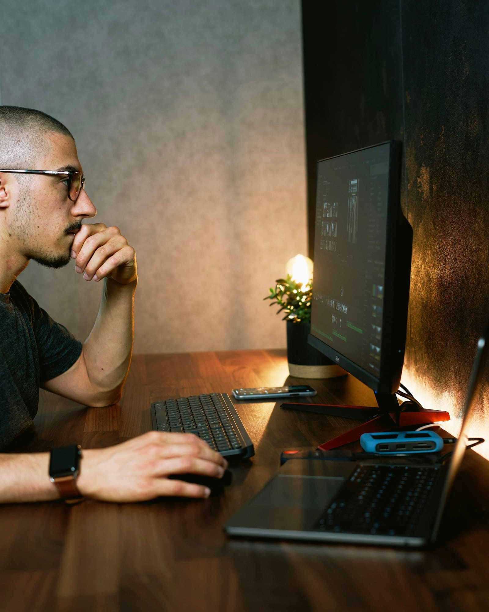 A man in glasses thinks at a wooden desk with a monitor, keyboard, laptop and a small warm lamp.