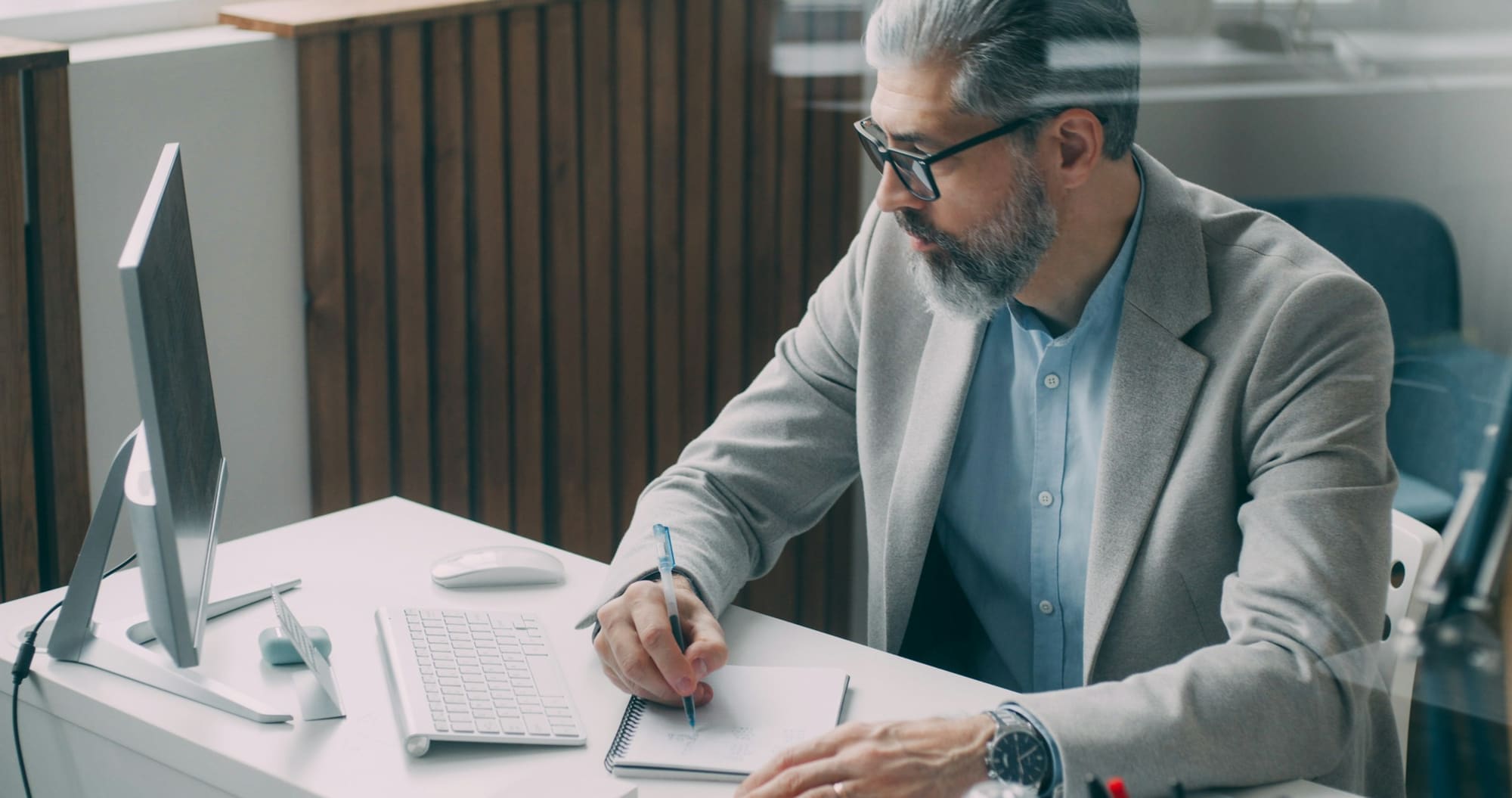 A man in a gray blazer writes notes at a bright desk beside a desktop computer.